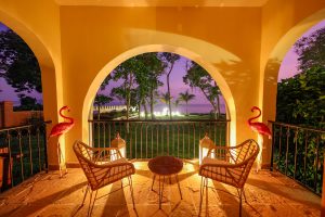 A stone terrace under three tall arches opens onto lawn and lake beneath a violet dusk sky, with cane chairs, glowing lanterns and two pink flamingo figures on either side.