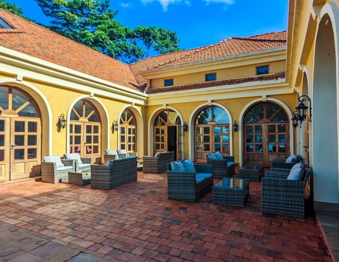 A brick-paved courtyard enclosed by yellow walls with arched glazed doors, furnished with grey wicker sofas and armchairs under a clear blue sky.