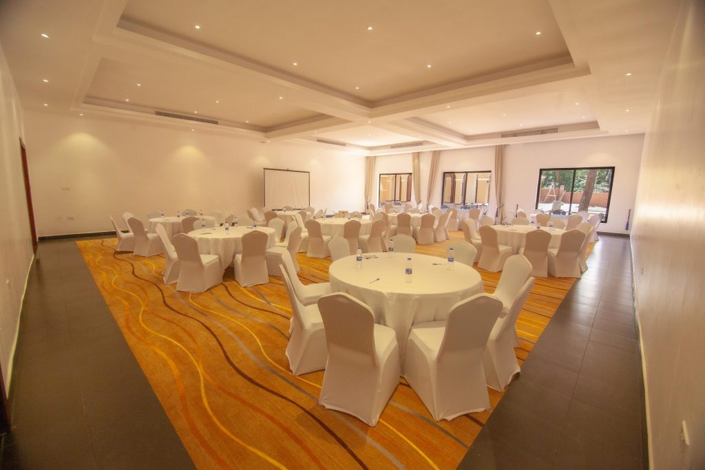 The same conference hall arranged for dining, with round tables in white linen and covered chairs on a patterned orange carpet.