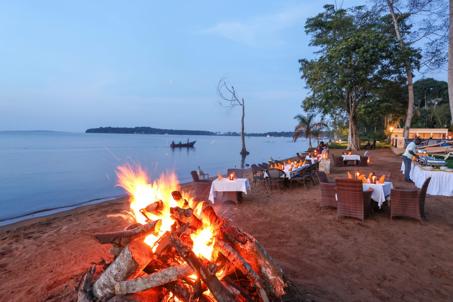 A large driftwood bonfire burns on a sand beach in front of white-clothed dining tables and wicker chairs, with a small boat on the calm lake beyond.