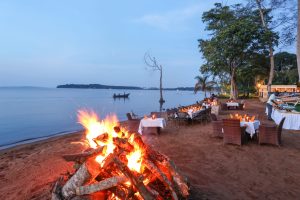 A large driftwood bonfire burns on a sand beach in front of white-clothed dining tables and wicker chairs, with a small boat on the calm lake beyond.