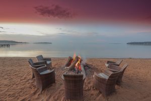 A log fire burning in a sand firepit ringed by wicker chairs on the beach, under a pink dusk sky over the lake.