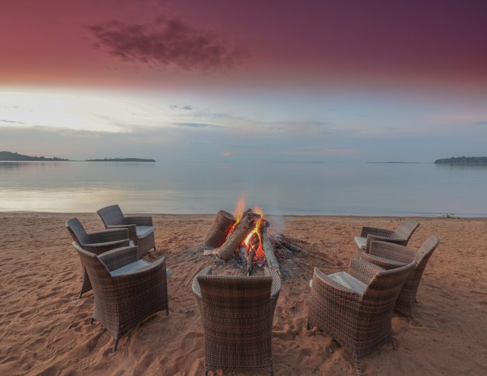 A log fire burning in a sand firepit ringed by wicker chairs on the beach, under a pink dusk sky over the lake.