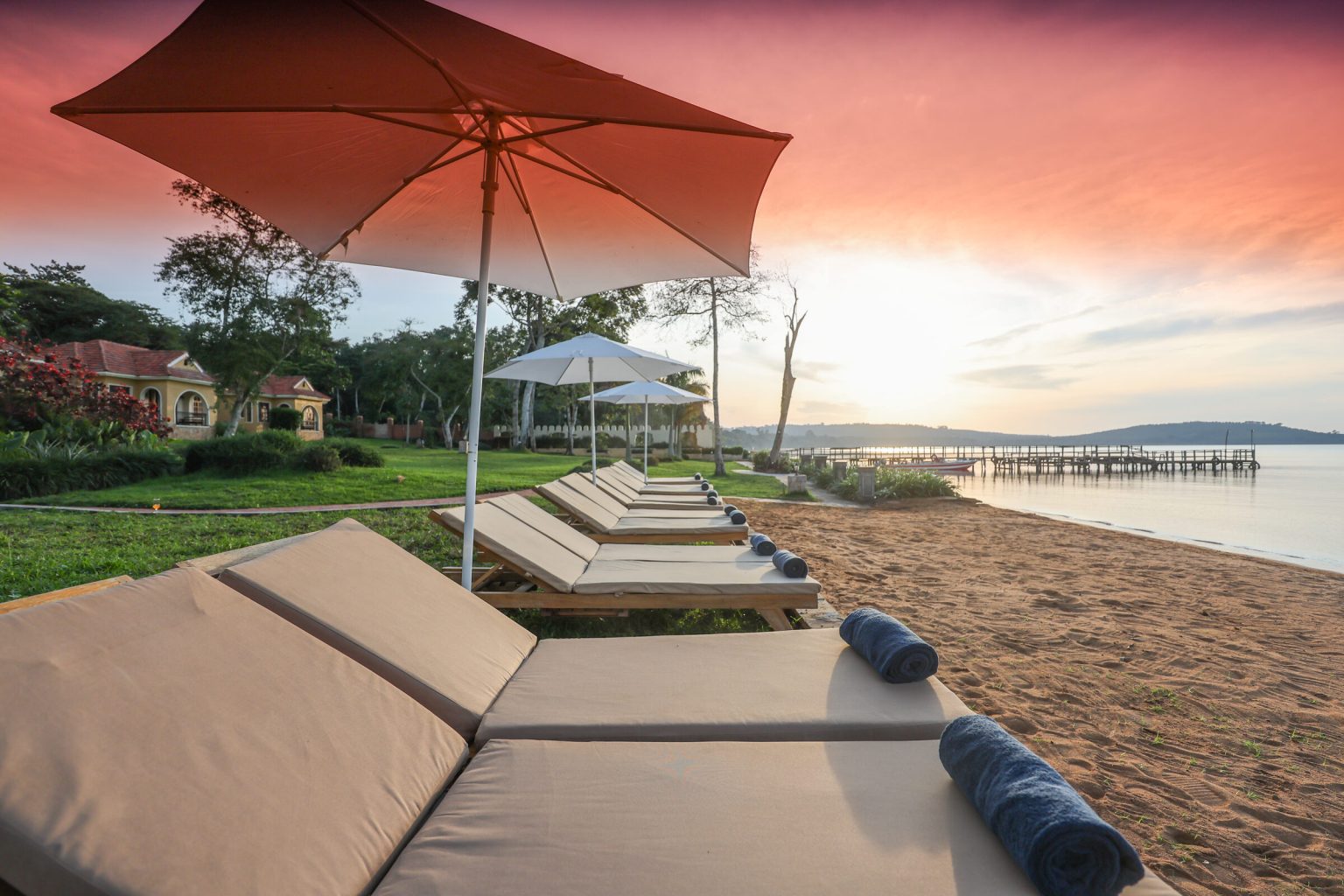 A row of cushioned sun loungers with rolled navy towels sits under parasols on a sand beach, facing a jetty and a pink and orange sunset over the lake.
