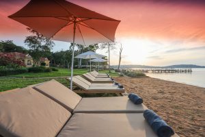 A row of cushioned sun loungers with rolled navy towels sits under parasols on a sand beach, facing a jetty and a pink and orange sunset over the lake.