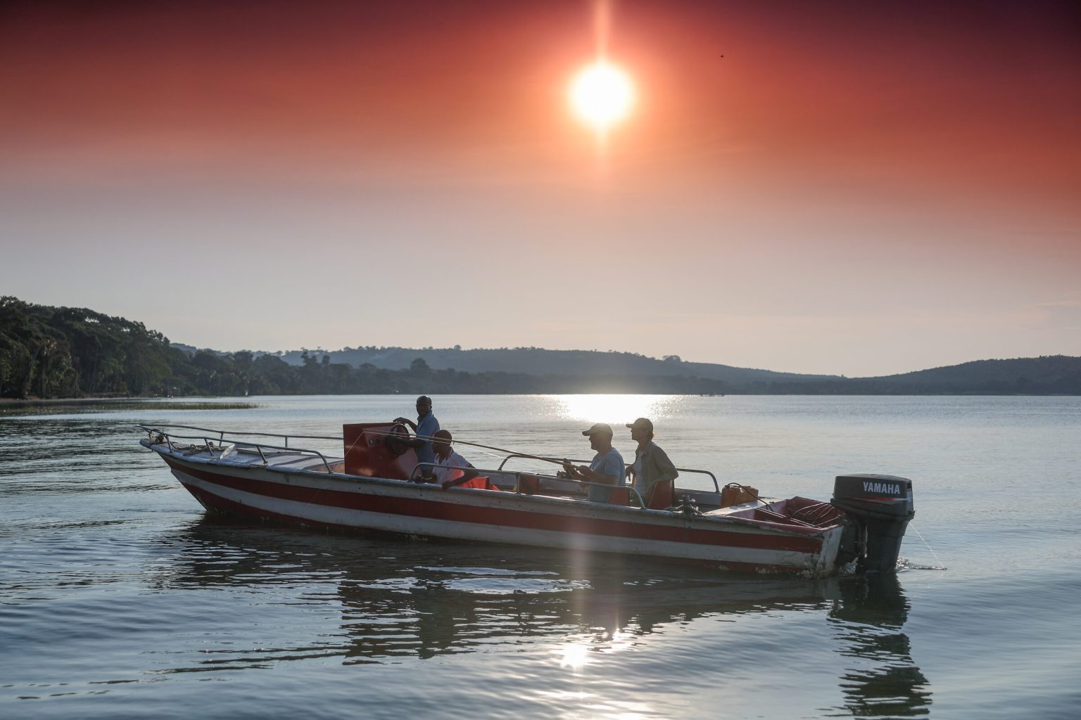 A motorboat carrying a small group sits on calm water beneath a low sun, its light spilling in a bright path across the lake.