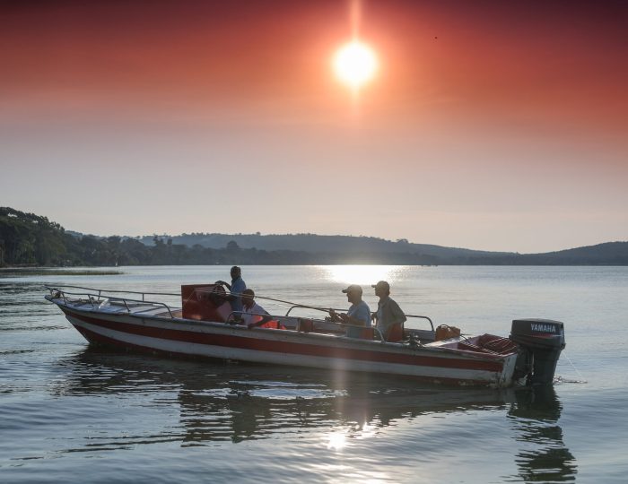 A motorboat carrying a small group sits on calm water beneath a low sun, its light spilling in a bright path across the lake.