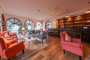 A bar lounge with grey and coral seating, a long bar of spirits and arched windows looking out to the lake.