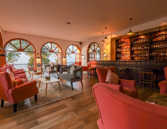 A warmly lit bar with arched windows, coral armchairs and shelves of spirits behind a dark wooden counter.