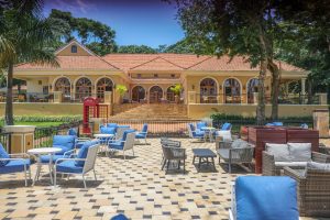 A long arcaded lodge building with a terracotta tile roof sits behind a large paved terrace of blue-cushioned outdoor furniture, under a clear blue sky.