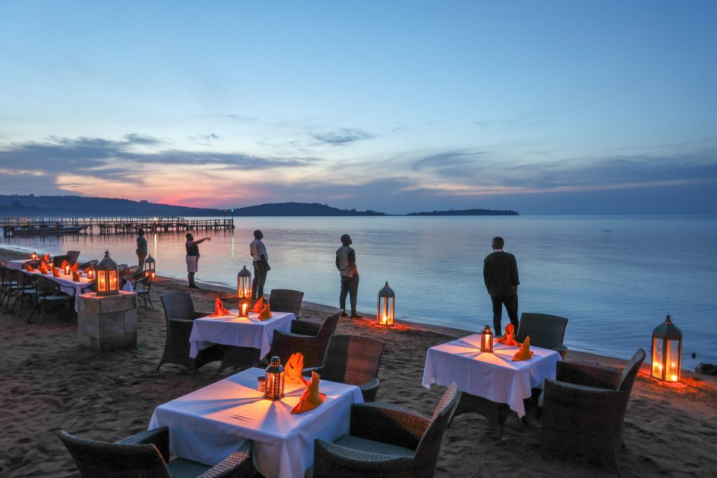 Tables laid with white linen and folded orange napkins are arranged on a sand beach among glowing lanterns, with figures silhouetted against a pink and blue twilight sky over the lake.