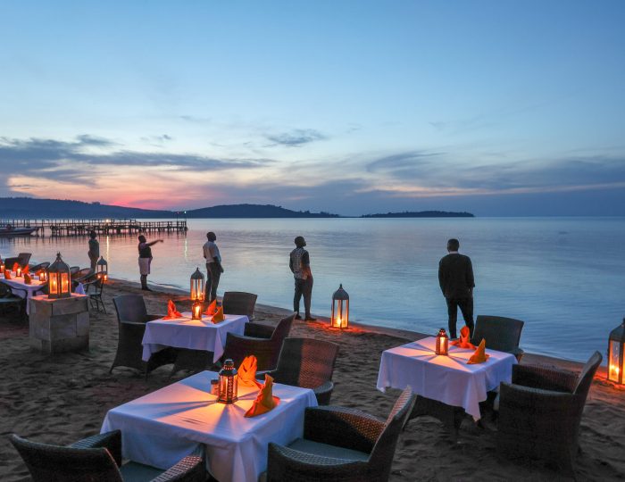 Tables laid with white linen and folded orange napkins are arranged on a sand beach among glowing lanterns.