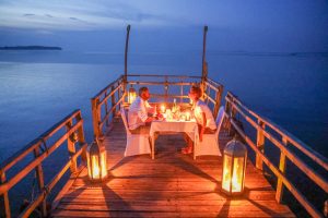 Seen from above, a couple dine at a small candlelit table at the end of a wooden jetty, framed by lanterns and the darkening lake.