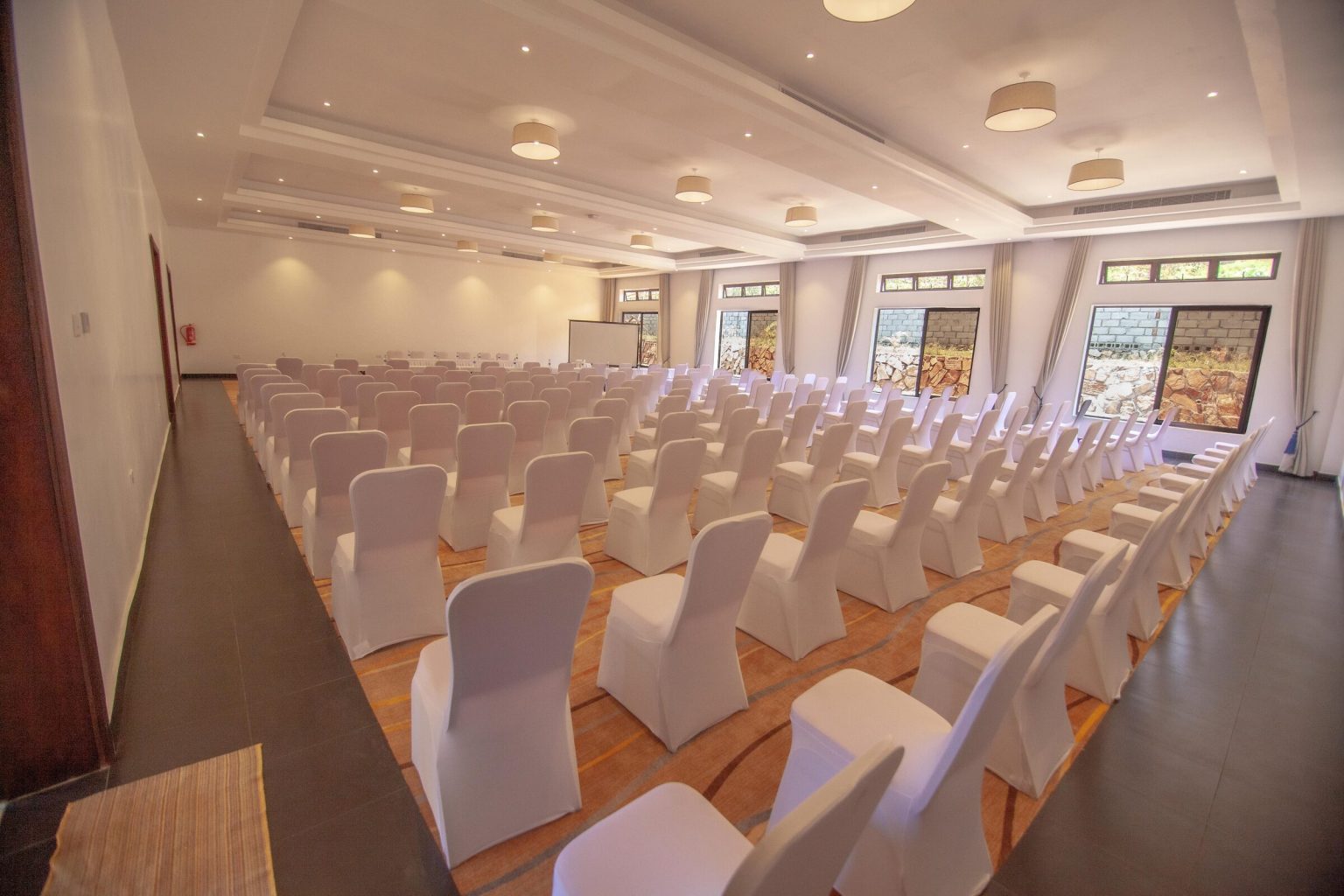 A large conference hall filled with rows of white-covered chairs facing a projector screen, lit by tall windows along one wall.