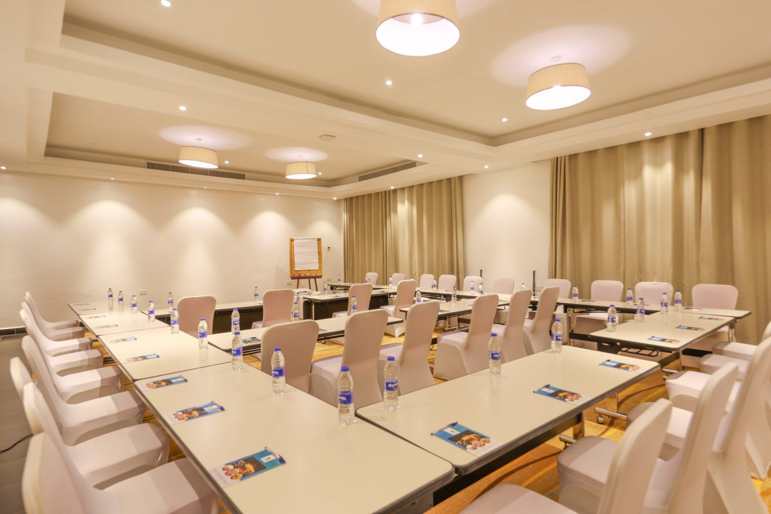 A conference room set with tables in a U-shape, white chair covers, bottled water and a flip chart at the front.