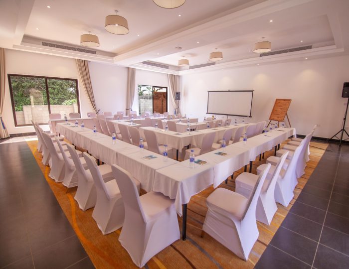A meeting room with tables in a U-shape under white linen and chair covers, set with notepads and water bottles, facing a pull-down projector screen and flipchart.