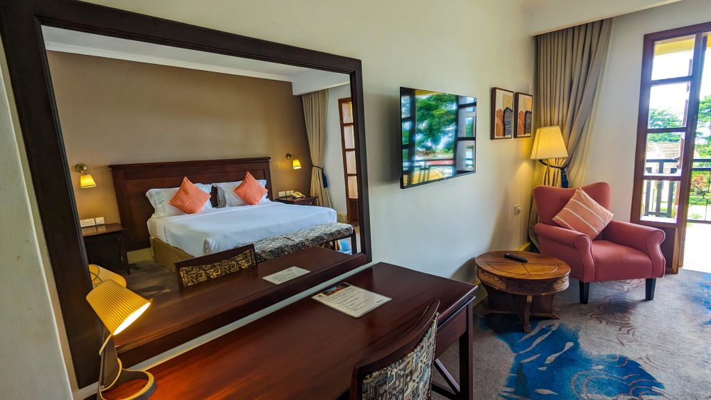 A guest room seen across a writing desk, with a large mirror reflecting a double bed with orange cushions, and a red armchair beside open balcony doors.