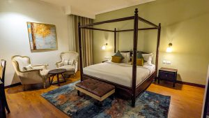 A bedroom with a dark wooden four-poster bed, a leather ottoman at its foot, two cream armchairs and a patterned rug on a polished timber floor.