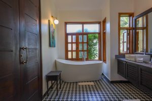 A freestanding bath set beneath an open window looking out to greenery, on a black and white chequered floor.