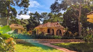 A brick path curves across a lawn towards yellow cottages with terracotta roofs, surrounded by dense green trees and flowering shrubs.