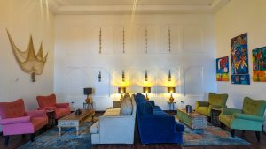 A tall, light lounge furnished with pink, green and blue armchairs and sofas around low tables, with a decorative fishing net and lanterns mounted on the white wall.