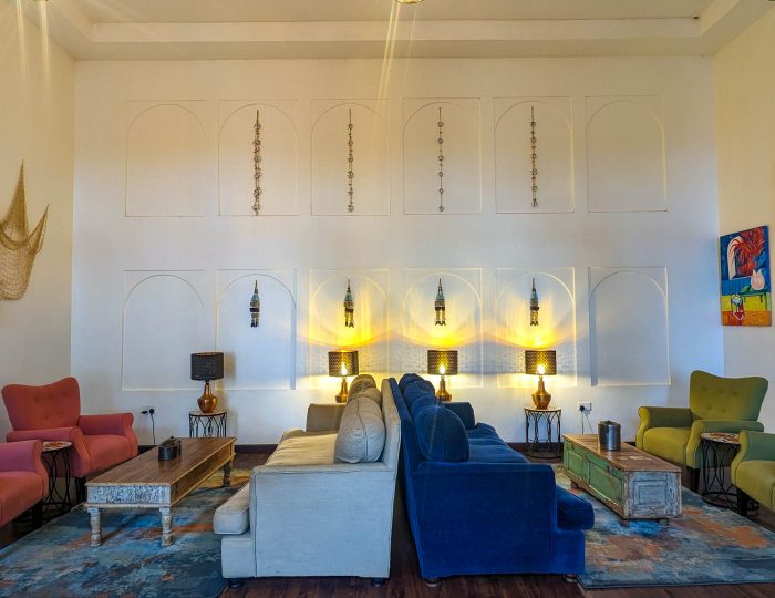 A tall, light lounge furnished with pink, green and blue armchairs and sofas around low tables, with a decorative fishing net and lanterns mounted on the white wall.