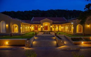 The lodge's arcaded courtyard glows with warm uplighting at dusk, its symmetrical wings framing a lawn and central water feature beneath a deep blue sky.