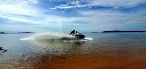 A jet ski throws up a wall of spray as it speeds across the lake just off a shallow sandy shore, with wooded islands on the horizon.