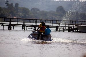 Two people in life jackets ride a jet ski across the lake in front of a long wooden jetty, throwing a fan of spray behind them.