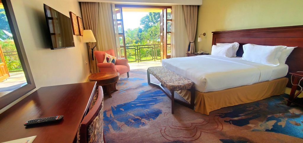 A hotel bedroom with a king bed, wall-mounted television, writing desk and an orange armchair beside open doors leading onto a balcony with a green garden view.