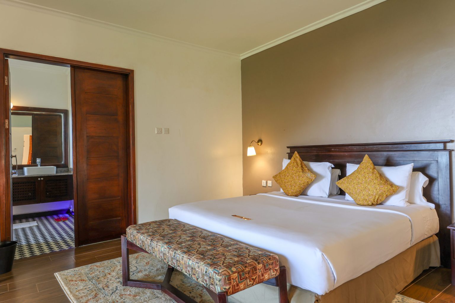 A king bed with gold cushions and an upholstered bench at its foot, next to a sliding door into the bathroom.