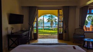 A guest room looking out through open arched doors to a balcony, a palm tree and the blue expanse of the lake beyond.