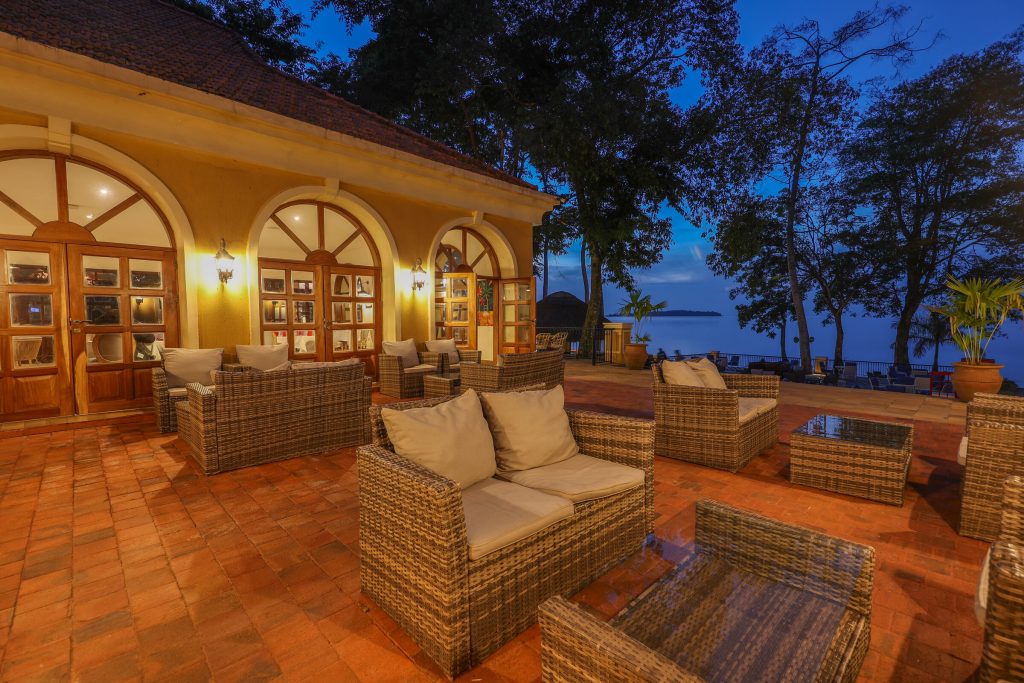 A brick terrace of wicker sofas outside lit arched doors at blue hour, with the lake and trees beyond.