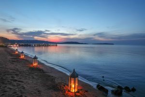 A line of glowing metal lanterns is set along the water's edge on a sand beach, leading the eye towards a jetty and a sunset over the lake.