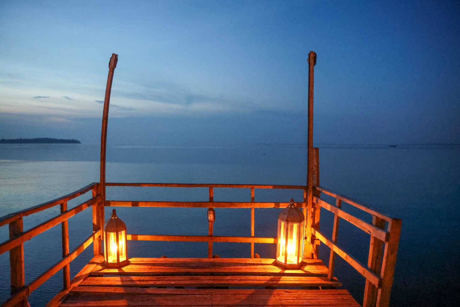 The end of a rustic wooden jetty at blue hour, with two glass lanterns burning on the deck and still water reaching to the horizon.