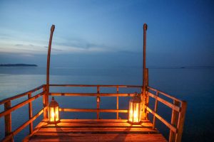 The end of a rustic wooden jetty at blue hour, with two glass lanterns burning on the deck and still water reaching to the horizon.