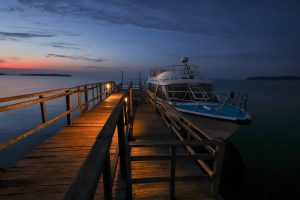 A wooden jetty lit by warm lamps leads out over the lake at twilight, with a motor boat moored at its far end and the last orange of sunset on the horizon.