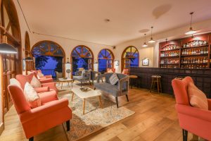 The lodge bar in the evening, with lamplit coral armchairs and arched windows showing deep blue night outside.