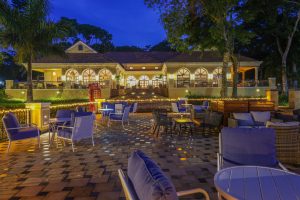 An arcaded lodge building glows with warm light above a paved terrace furnished with blue lounge chairs and tables, strung with fairy lights under a deep blue night sky.