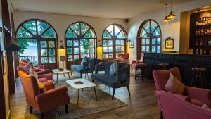 A lounge bar with a dark timber counter and stools stocked with spirits, opening onto a seating area of coral and blue armchairs beside tall arched lake-facing windows.