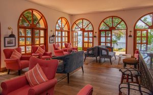 A lounge of coral wingback armchairs and grey sofas faces a row of tall arched French doors that open onto a terrace with the lake beyond.