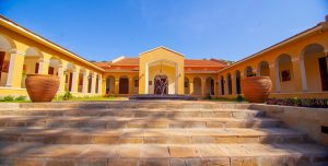 Broad stone steps lead up between two large terracotta urns to a yellow arcaded building with a small fountain playing in front of its central arched entrance.