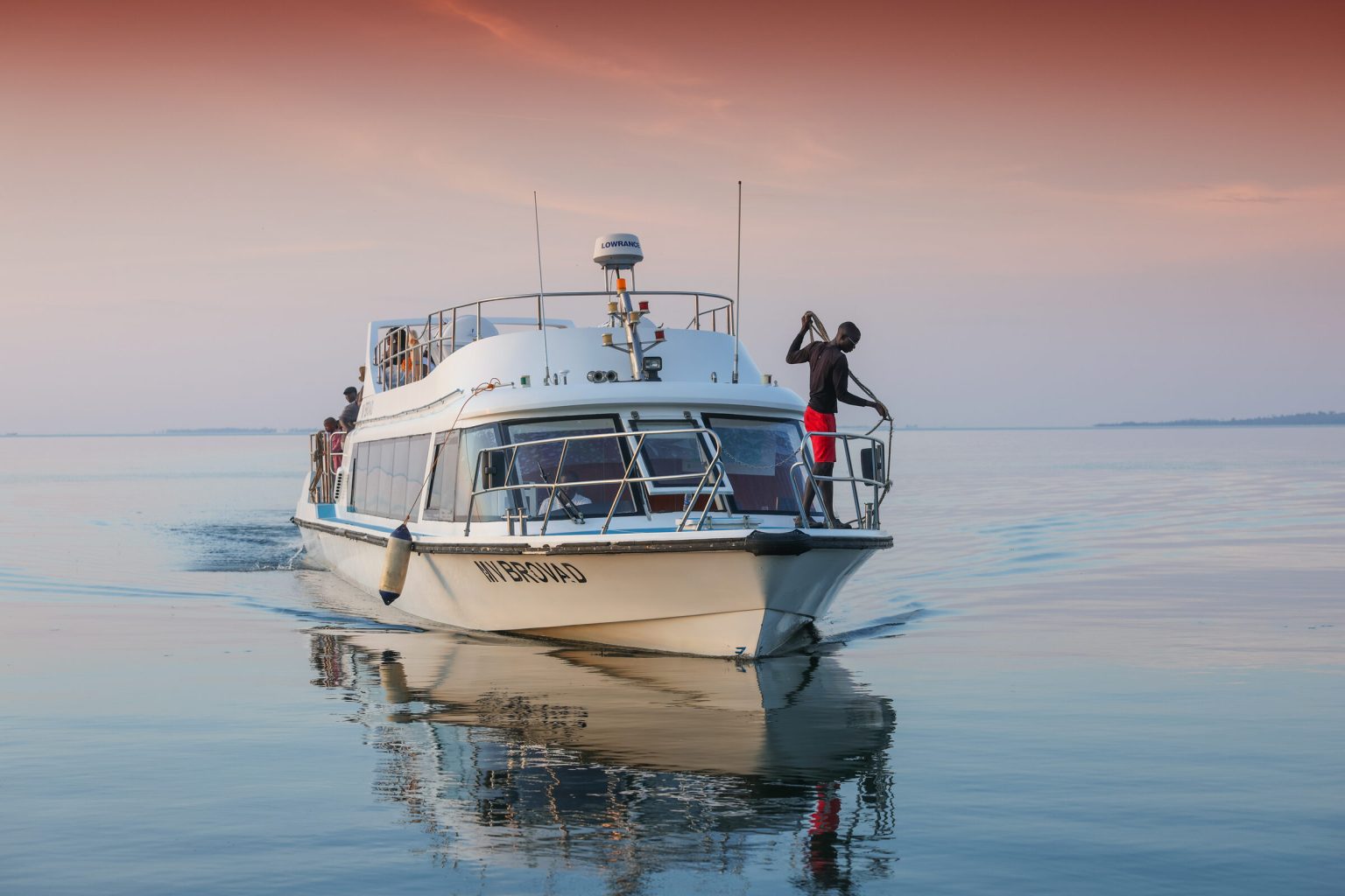 The MV Brovad motor boat glides across still, pink-lit water with a crew member standing at the bow.