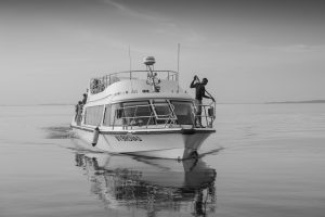 A white passenger boat named MV Brovad moves across glassy open water, its reflection mirrored beneath it, with a crew member standing at the bow.