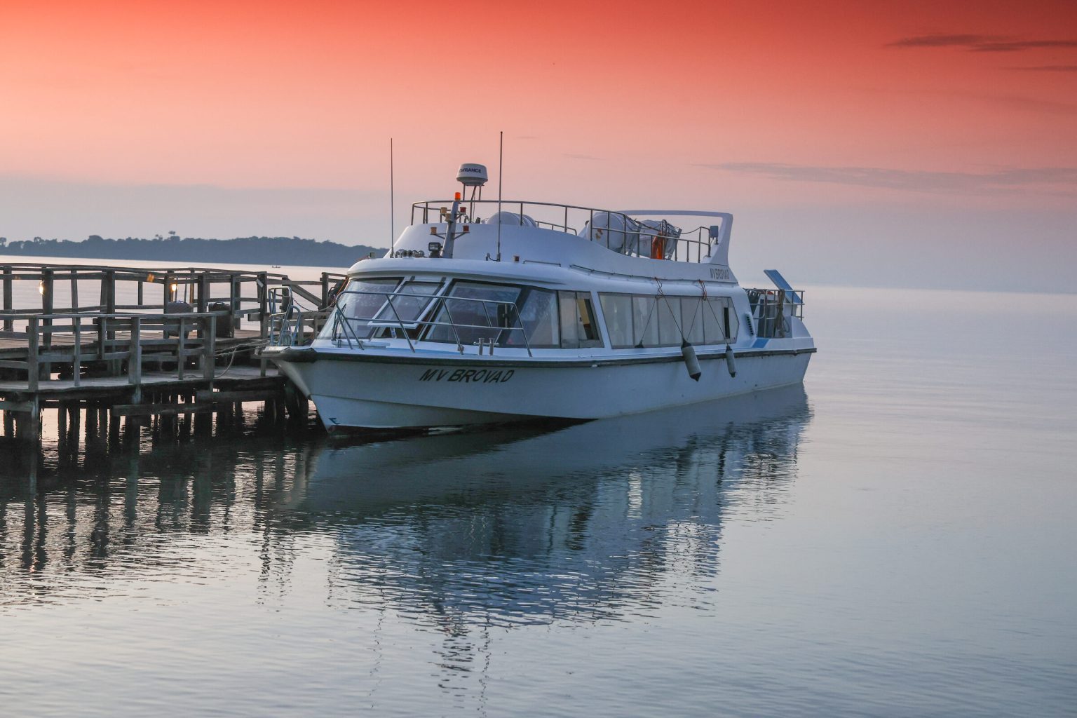 The MV Brovad sits moored alongside a wooden jetty on glassy water beneath a red and pink sky.