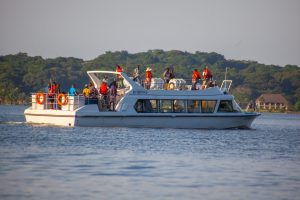 The passenger boat MV Brovad cruises past a green forested shoreline with a crowd of passengers standing on its open upper deck.