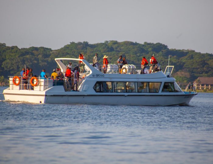 The passenger boat MV Brovad cruises past a green forested shoreline with a crowd of passengers standing on its open upper deck.