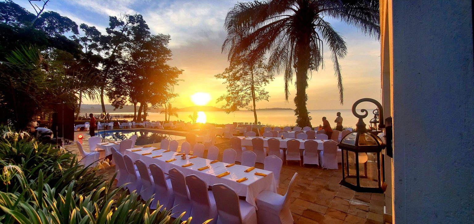 Long banquet tables in white linen and chair covers are laid beside the pool as the sun sets over the lake behind silhouetted palms.