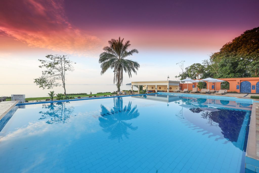 A poolside view at sunset with a palm reflected in the water and the lodge's poolside bar and rooms to one side.