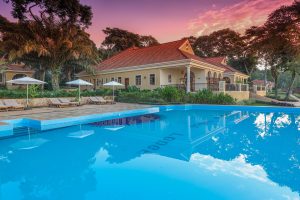 Sun loungers and white umbrellas beside a swimming pool, with the tile-roofed lodge building behind under a pink sky.